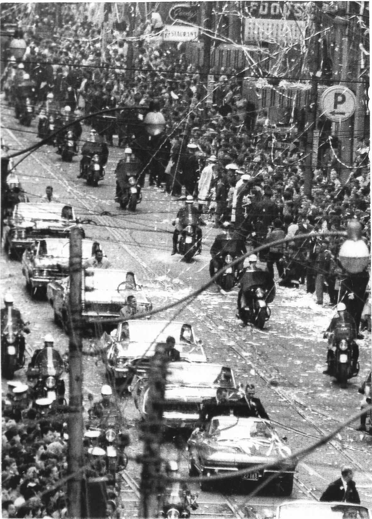A black-and-white photograph of a crowded downtown street during a parade, confetti and streamers in the air, motorcycle escorts and open convertible cars moving through the crowd, overhead wires crossing the frame. Bay Street, Toronto, 1967.